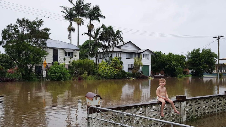 A little boy smiling as he sits on a front fence in a house in a flooded street