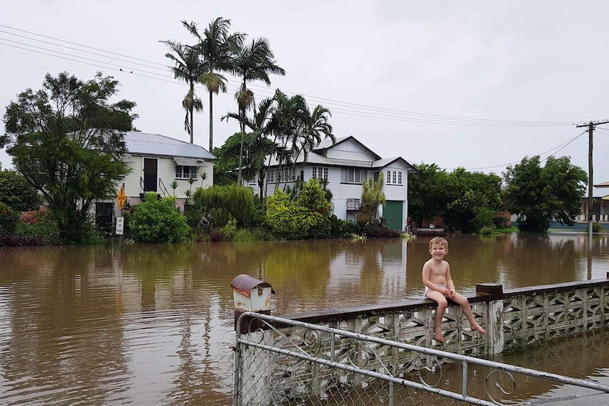 A little boy smiling as he sits on a front fence in a house in a flooded street