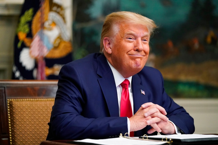 You see Donald Trump, wearing a blue suit and red tie, smiling as he sits at a desk.