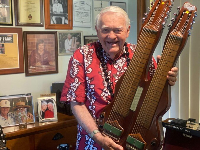 Photo of Kenny Kitching smiling widely. He has white hair and wears colourful print shirt, and holds a guitar with two necks.