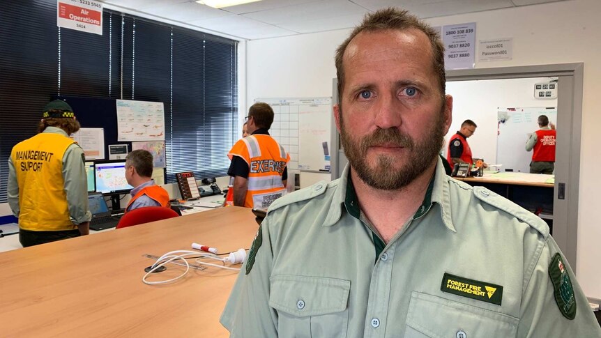 A man in a green Forest Fire Management uniform stands in an office as workers in uniform talk together in the background.