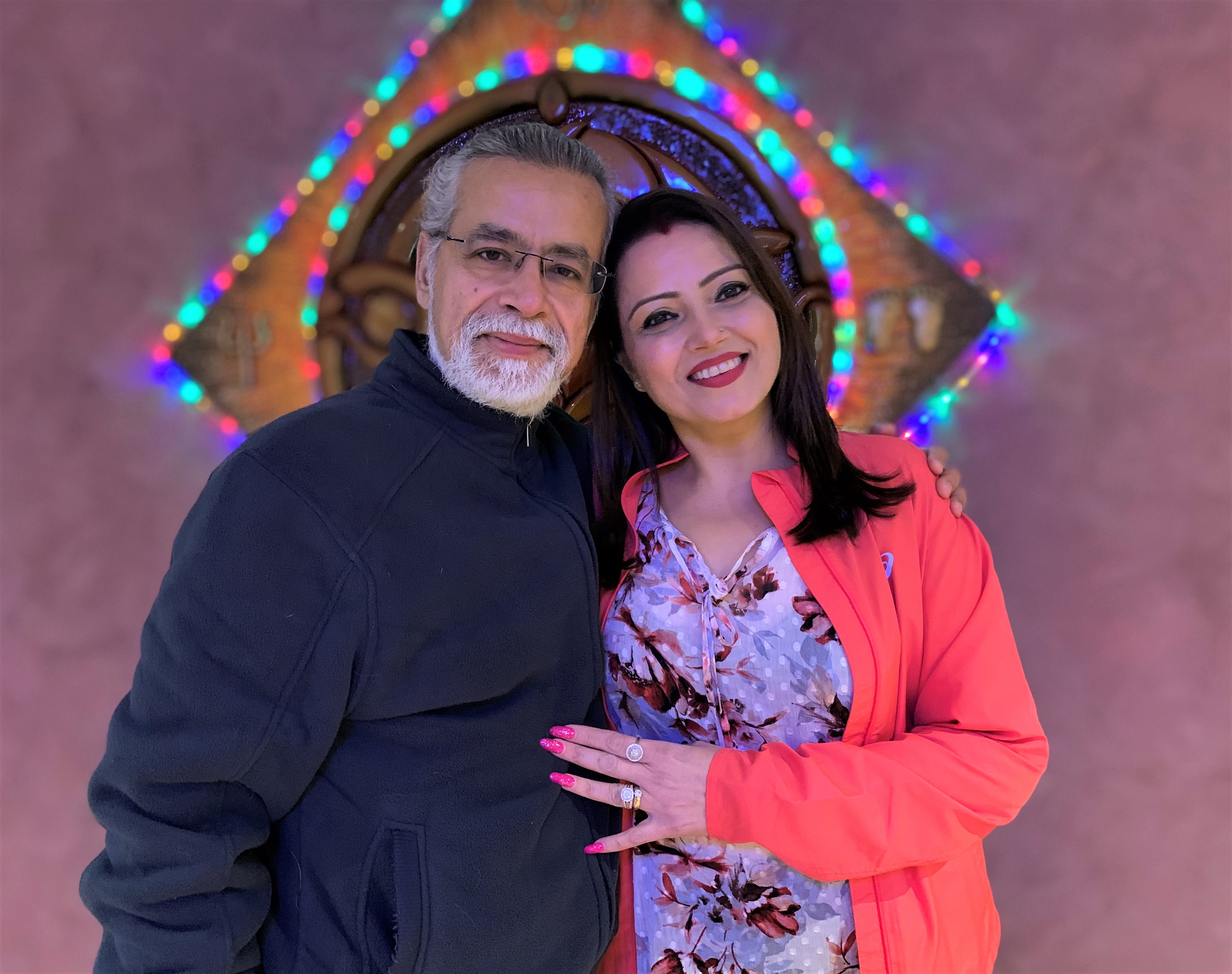 A man and a woman cuddle in front of some sparkly lights at their home