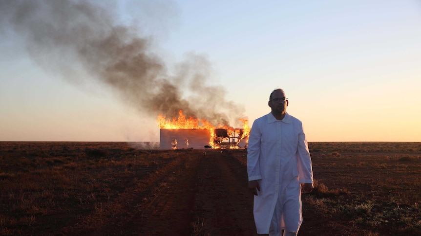 A man dressed in white walks toward the camera, in the background a wooden wall on a red dirt road burns.