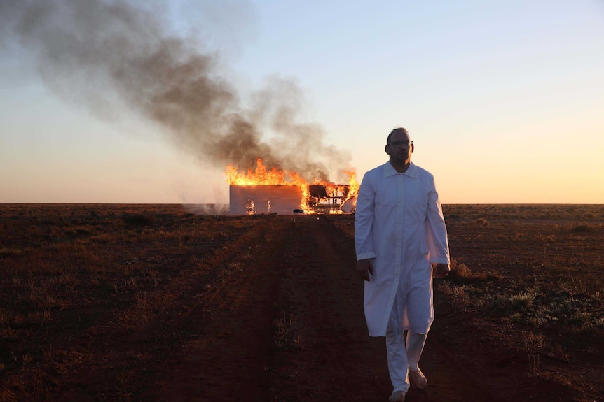 A man dressed in white walks toward the camera, in the background a wooden wall on a red dirt road burns.