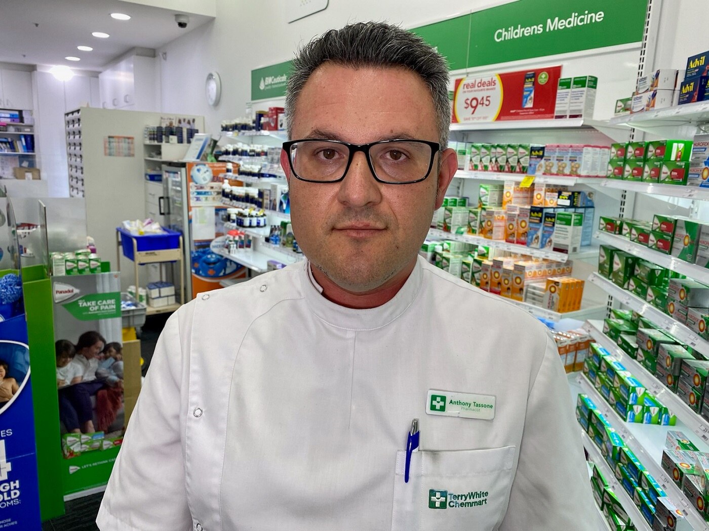 Anthony Tassone stands inside a pharmacy, dressed in a white coat with a name badge.