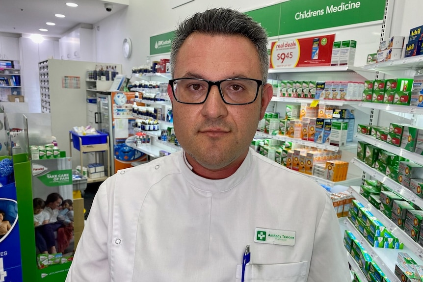Anthony Tassone stands inside a pharmacy, dressed in a white coat with a name badge.
