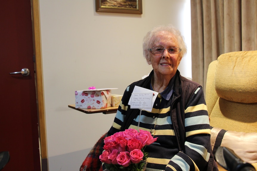 An older woman with white hair and glasses grins at the camera, sitting with pink roses in her lap
