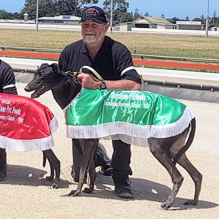 A man in a black shirt and cap crouches down next to a racing greyhound with a silk coat at a race track.