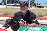 A man in a black shirt and cap crouches down next to a racing greyhound with a silk coat at a race track.
