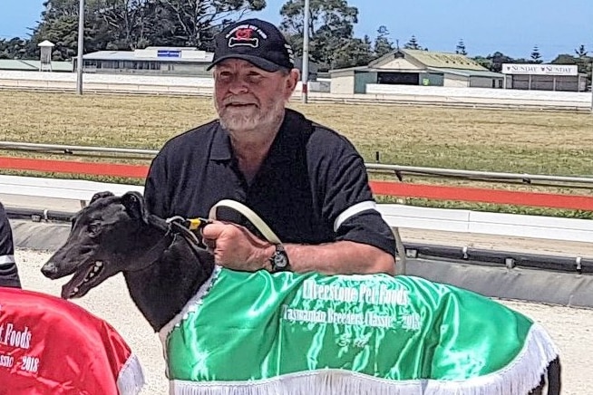 A man in a black shirt and cap crouches down next to a racing greyhound with a silk coat at a race track.