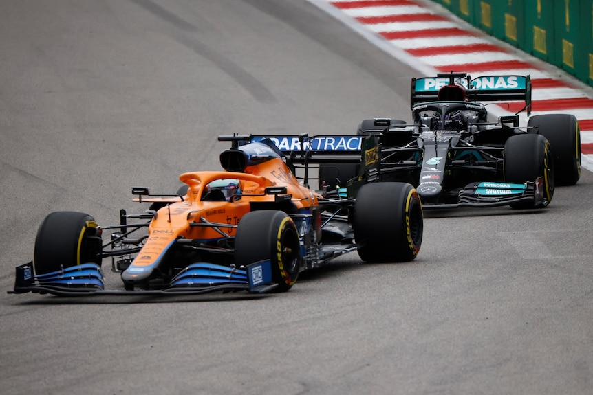 Un coche de fórmula uno naranja seguido de un coche turquesa en una pista