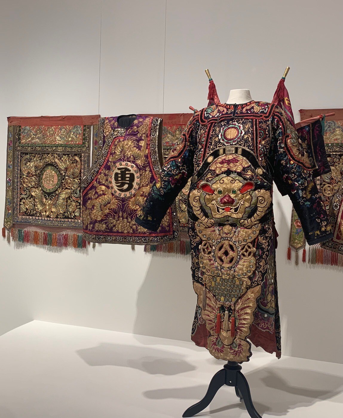 A long elaborate coat with flags attached to the shoulders posed on a stand, next to a similar waistcoat and several banners.