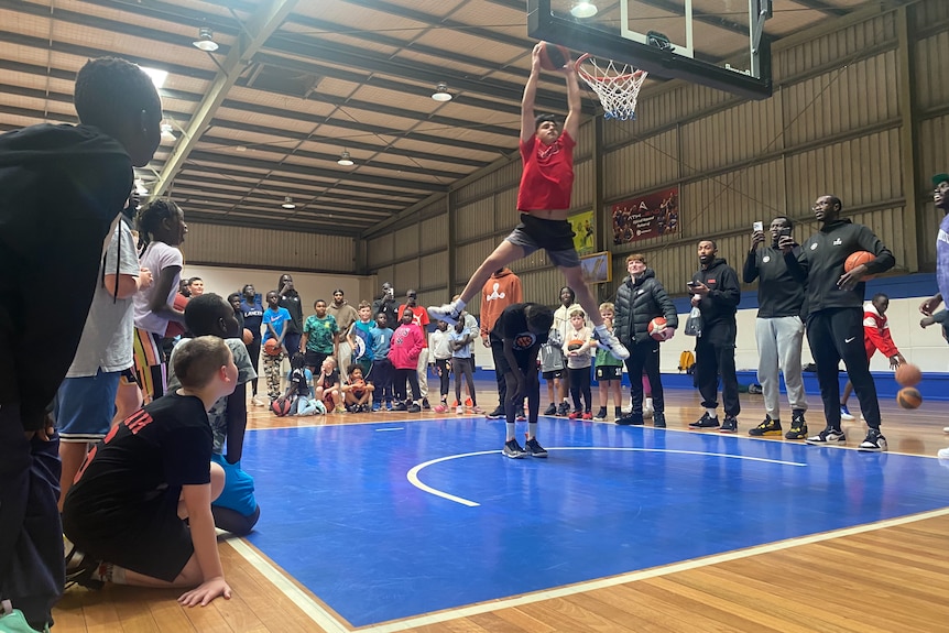 A group of people crowded around a basketball hoop with a boy jumping high with the ball. 