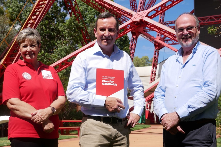 A woman and two men smiling at the camera, pictured in front of a metal structure/mining head frame.