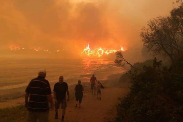 People walk down a beach, while flames rise in the distance, the sky is orange.