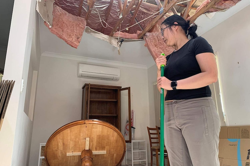 Sophia Wei holds a broom in a room in her hail-damaged home with smashed roof above her.