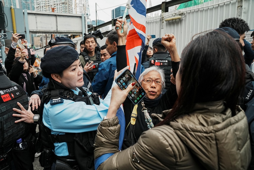 Several protesters gather outside a courthouse, one waving a British flag, while security forces attempt to stop the wave.