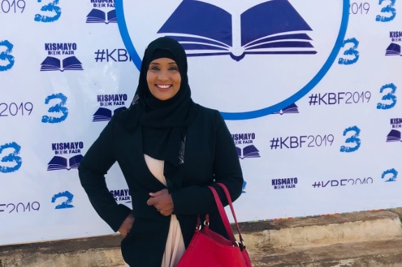 A woman in a black headscarf in front of a book festival sign holding a red handbag.