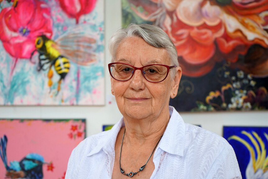 A lady with white hair and round glasses looks at the camera with a colourful background behind her.