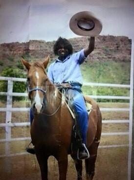 A picture of Mr Yeeda smiling on a horse, holding his hat in the air with Kimberley rock formations behind him