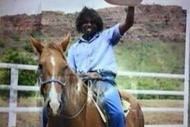 A picture of Mr Yeeda smiling on a horse, holding his hat in the air with Kimberley rock formations behind him