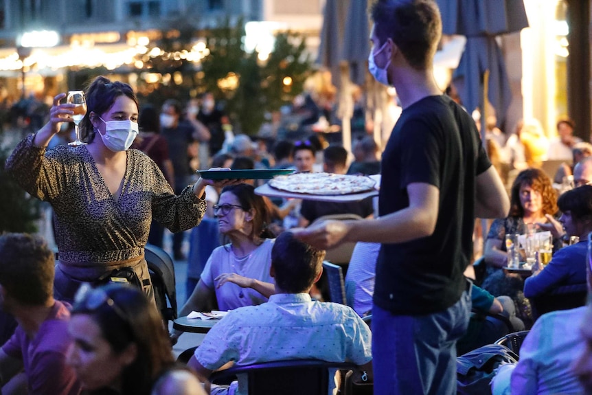 A man stands amongst a seated crowd holding a pizza. A woman by him has a glass of wine. Both are wearing surgical masks.