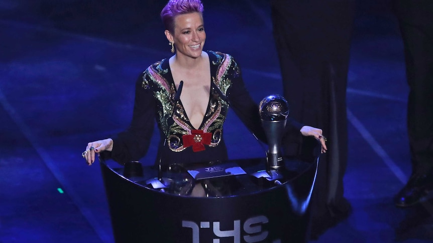 Megan Rapinoe smiles whilst standing at a podium with 'The Best' written on it