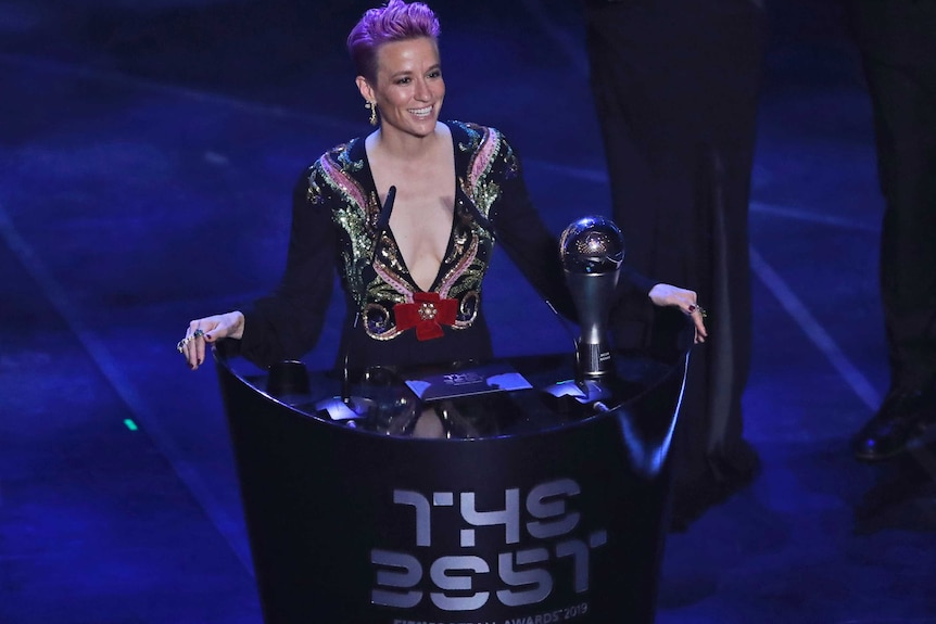 Megan Rapinoe smiles whilst standing at a podium with 'The Best' written on it