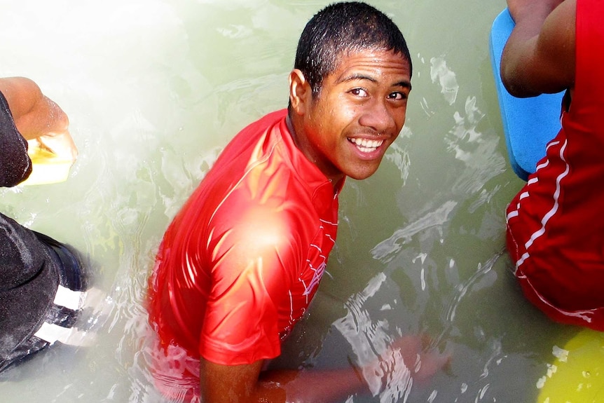 Finau is waist deep in water and wearing a red swim shirt. He looks upward smiling at the camera.