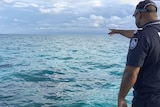 A police officer stands pointing out at the ocean from a boat, snorkels from several divers poke through the waves.