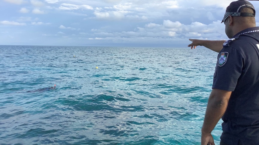 A police officer stands pointing out at the ocean from a boat, snorkels from several divers poke through the waves.