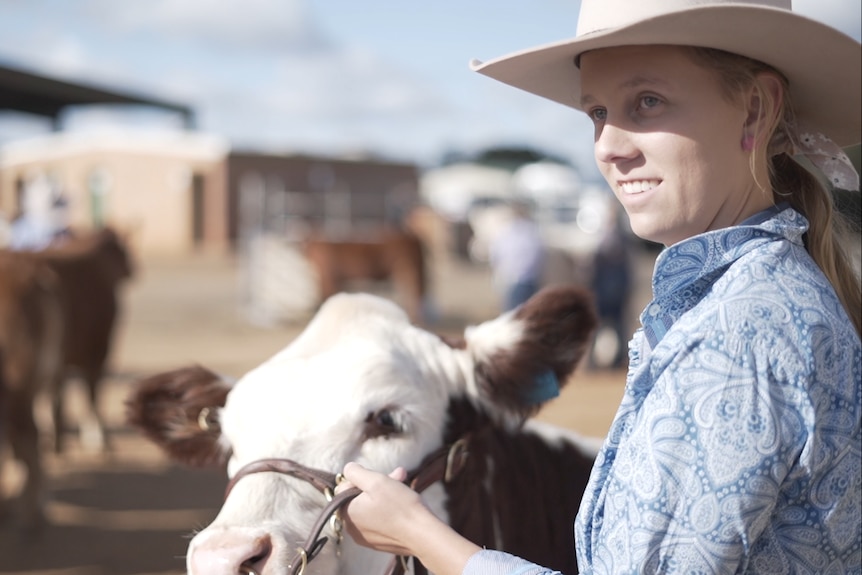 A young woman in a hat, holding a leashed cow.