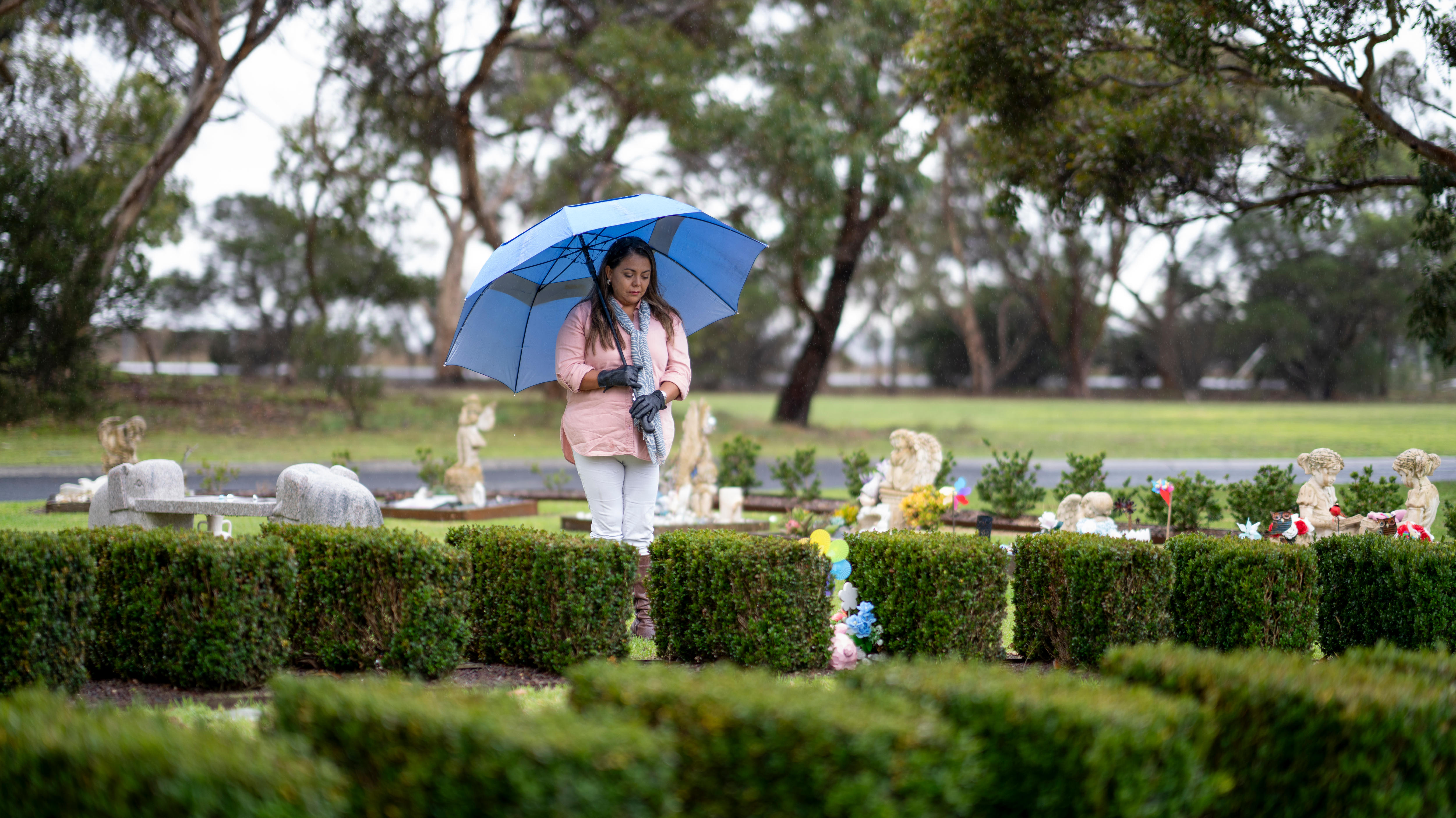 A woman wearing a pink top and white pants looks down at a grave holding a blue umbrella