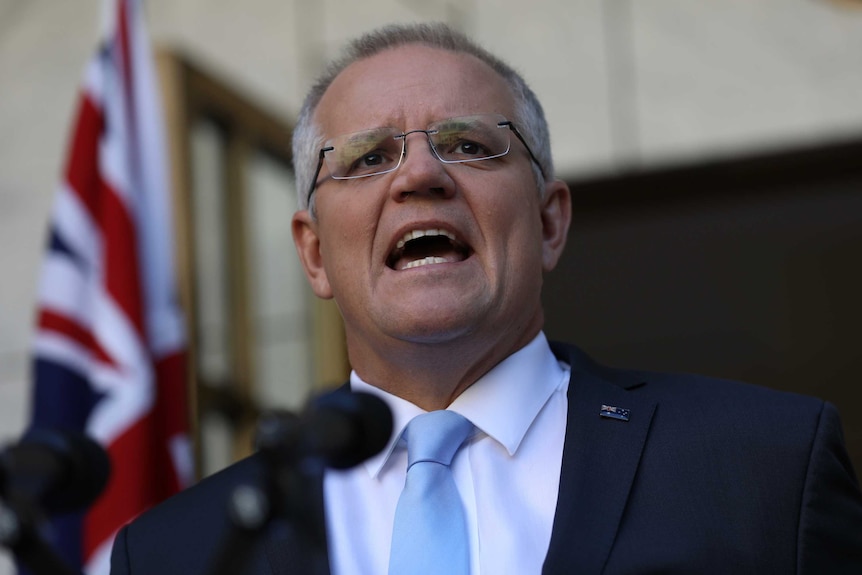 Wearing a black suit and a light blue tie, Scott Morrison speaks behind microphones.