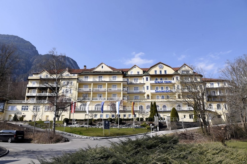 You view a German classical alpine hotel painted in pale yellow set against a blue sky with mountains in the distance.