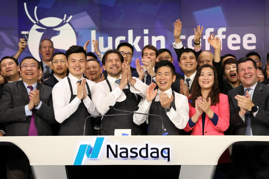 A group of Luckin employees and their CEO stands behind a desk with "Nasdaq" on it.  Some raise their hands, cheering