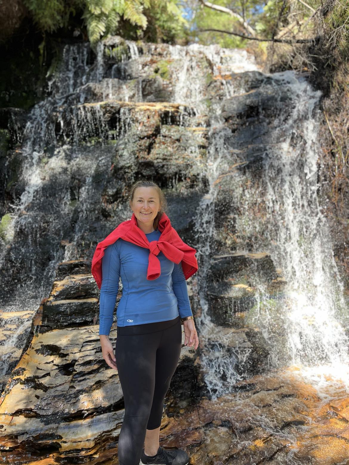 Alana standing in front of a waterfall. She's wearing a blue sweater, with a red jumper tied over her shoulders.