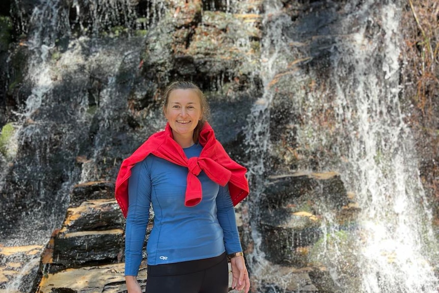 Alana standing in front of a waterfall. She's wearing a blue sweater, with a red jumper tied over her shoulders.