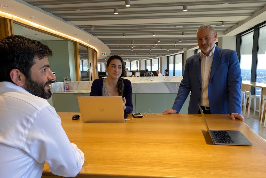 A grey-haired man, wearing a blue jacket, stands with two other workers who are sitting at a table chatting.