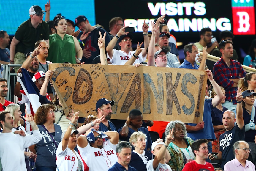 A large group of blue and white-clothed Yankees fans at a stadium, holding or standing near a large sign saying 'Go Yanks'.