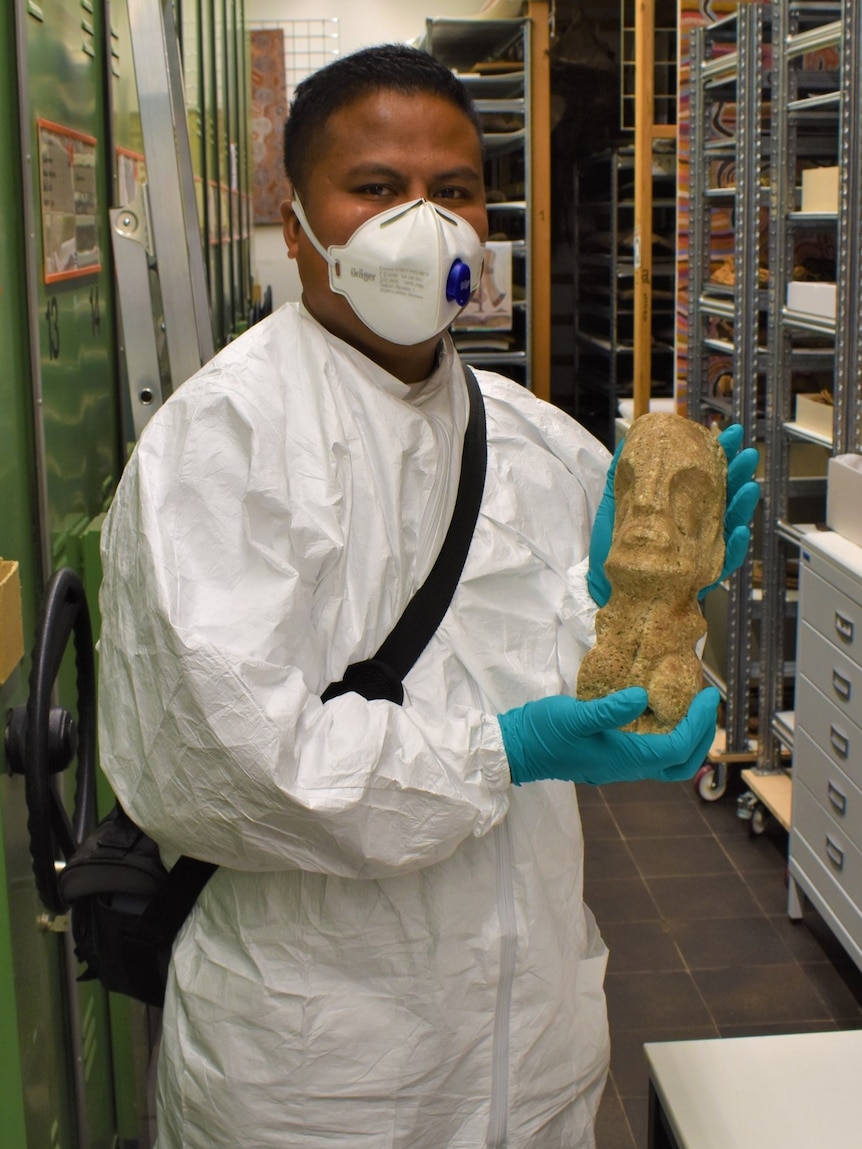 A man wearing a white gown and p2 mask holds up a yellow coloured carved Pacific Island artefact