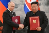Two men are shaking hands and holding folders while standing in front of country flags.