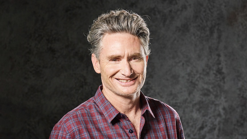 Comedian Dave Hughes smiles at camera while wearing a plaid shirt