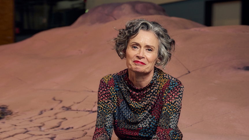 A woman sits on an articifial desert scene inside a theatre.