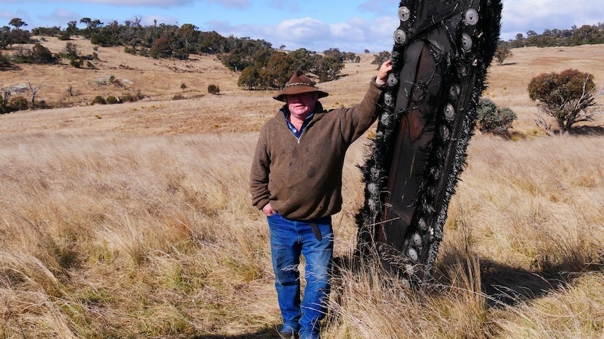 Man in wide brimmed hat stands in field leaning on a tree