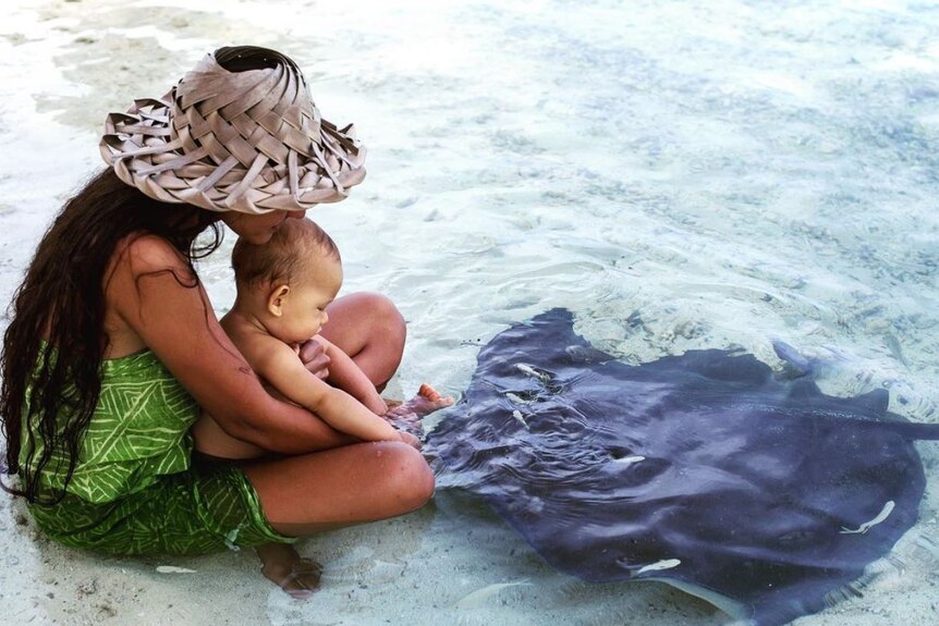 Woman wears handcrafted hat sitting in water on beach with baby patting a stingray