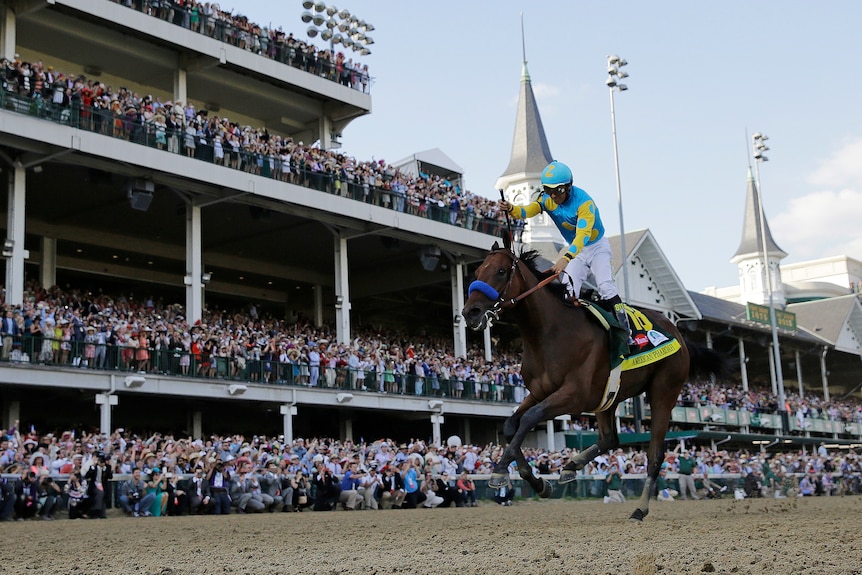 A jockey raises his fist in victory, riding a horse in the Kentucky Derby, in front of a massive grandstand of fans.