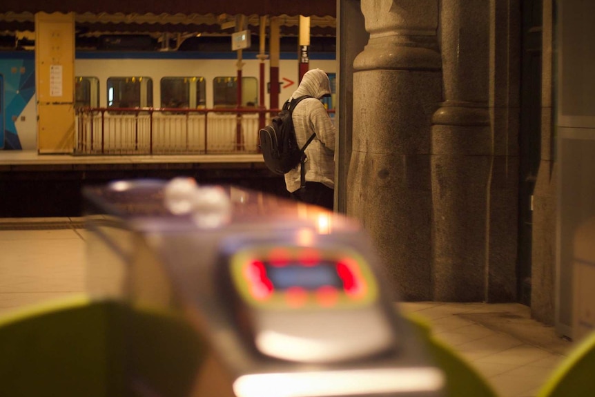 Behind the ticket barriers at Flinders Street Station, a man in a hoodie waits for a train on the platform.