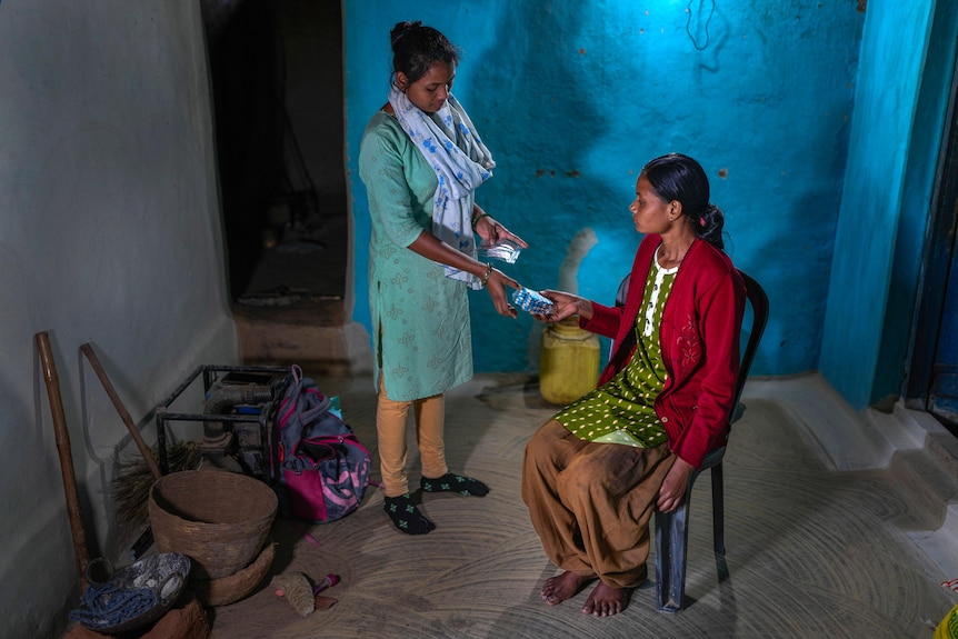 A woman hands medicine to a patient who is sitting in a chair in a room with blue and white walls
