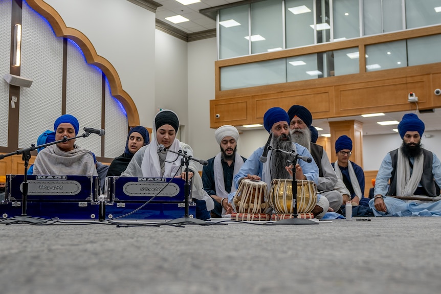 Men and women in turbans and traditional dress sit cross-legged and play drums and other instruments.